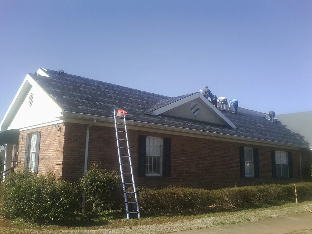 Crew tearing shingles off a brick ranch roof with a ladder set — Metal Roof Repair work by our crew in De Mossville