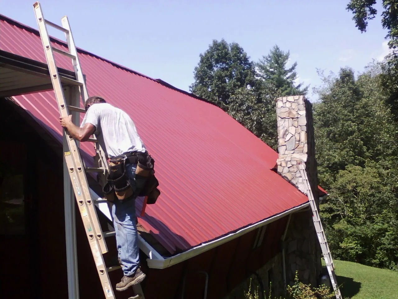 Roofer on a ladder beside a red metal roof and stone chimney — Soffit & Fascia Repair by Roofing Brothers in De Mossville